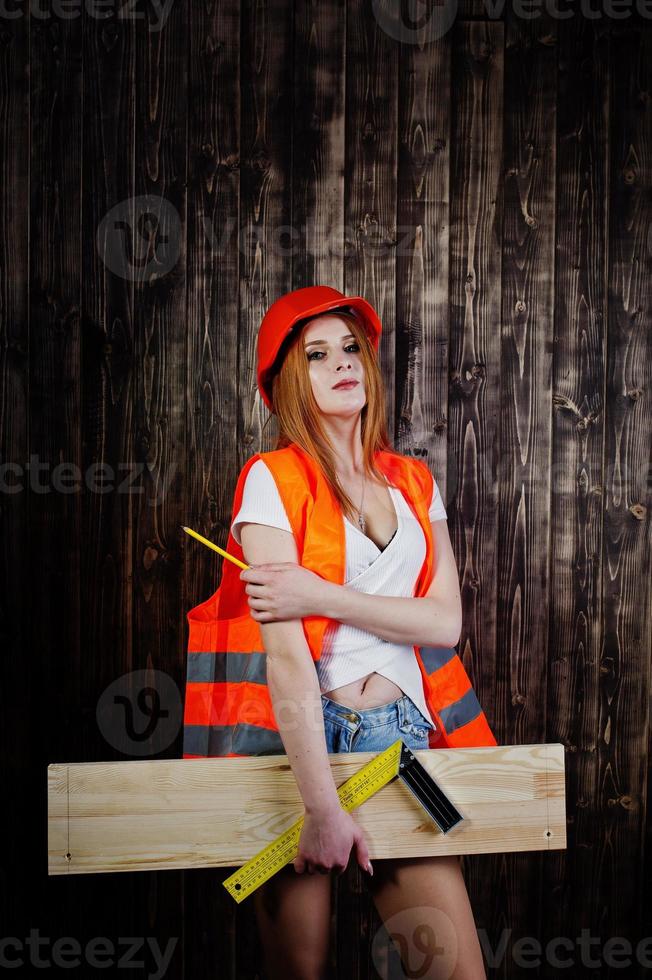 mulher engenheira em laranja protege o capacete e a jaqueta de construção contra o fundo de madeira, segurando a régua e a placa. foto