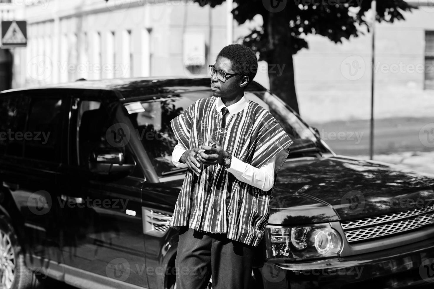 empresário africano em roupas tradicionais e óculos com celular contra carro preto suv. ricos africanos. foto