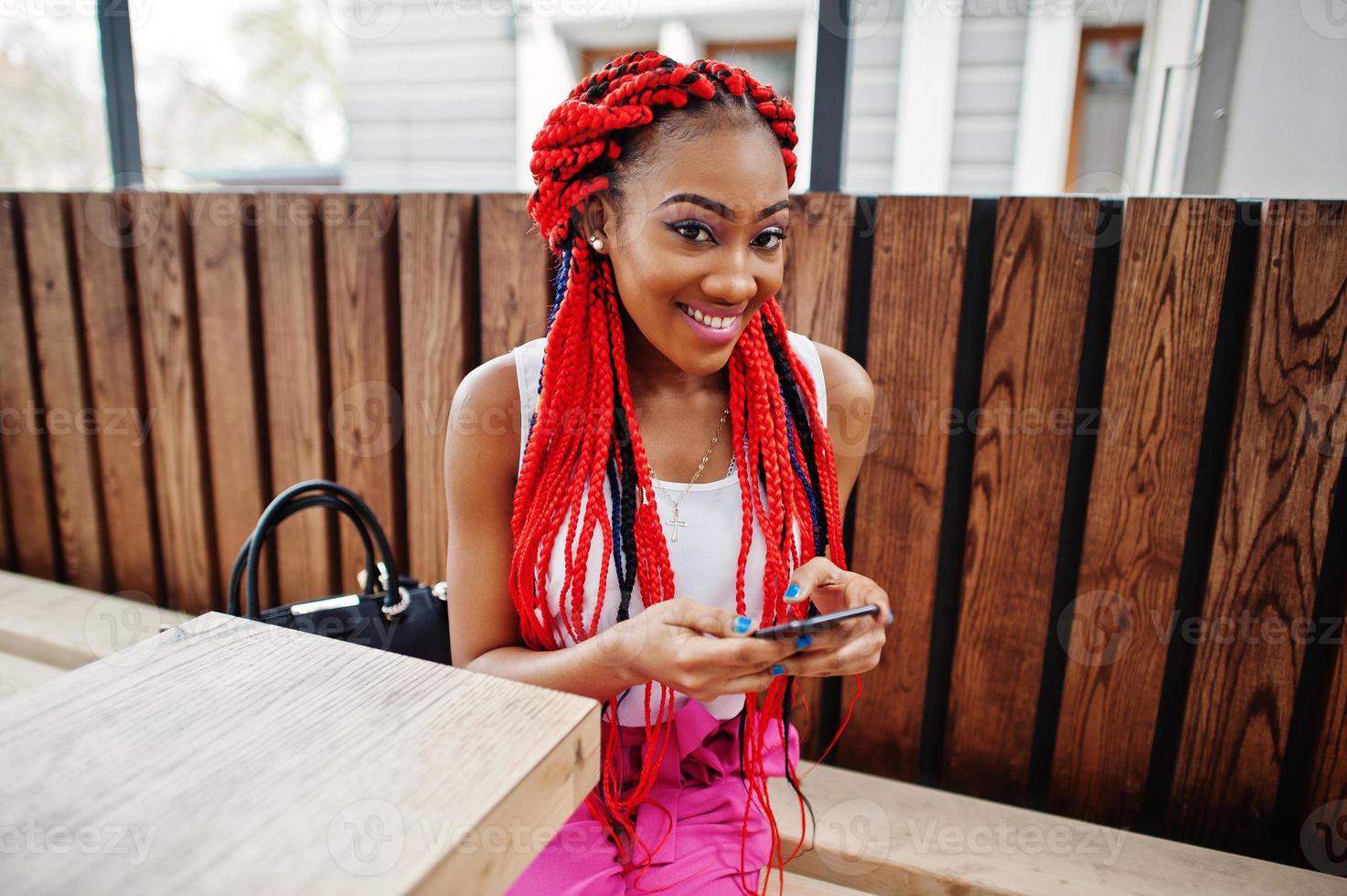 garota afro-americana na moda em calças cor de rosa e dreads vermelhos posou ao ar livre com telefone celular. foto