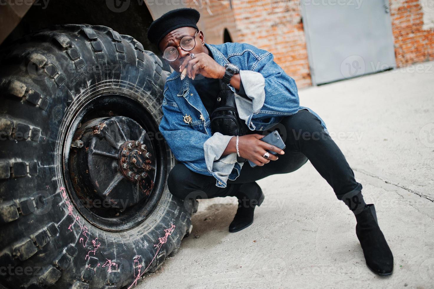 homem afro-americano de jaqueta jeans, boina e óculos, fumando charuto e posou contra a roda do veículo blindado militar btr. foto