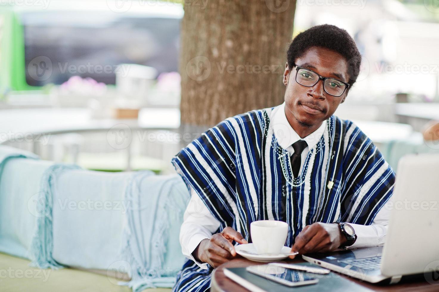 homem africano em roupas tradicionais e óculos sentados no caffe ao ar livre, bebendo café e trabalhando no laptop. foto