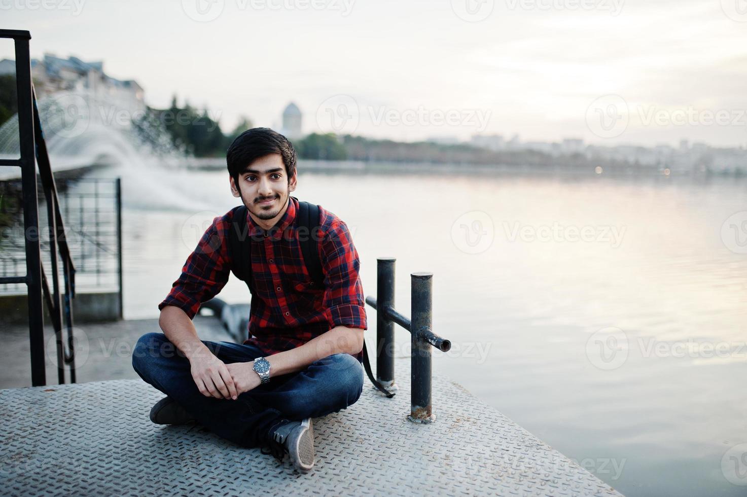 homem jovem estudante indiano na camisa quadriculada e jeans com mochila posou na cidade à noite contra fontes. foto