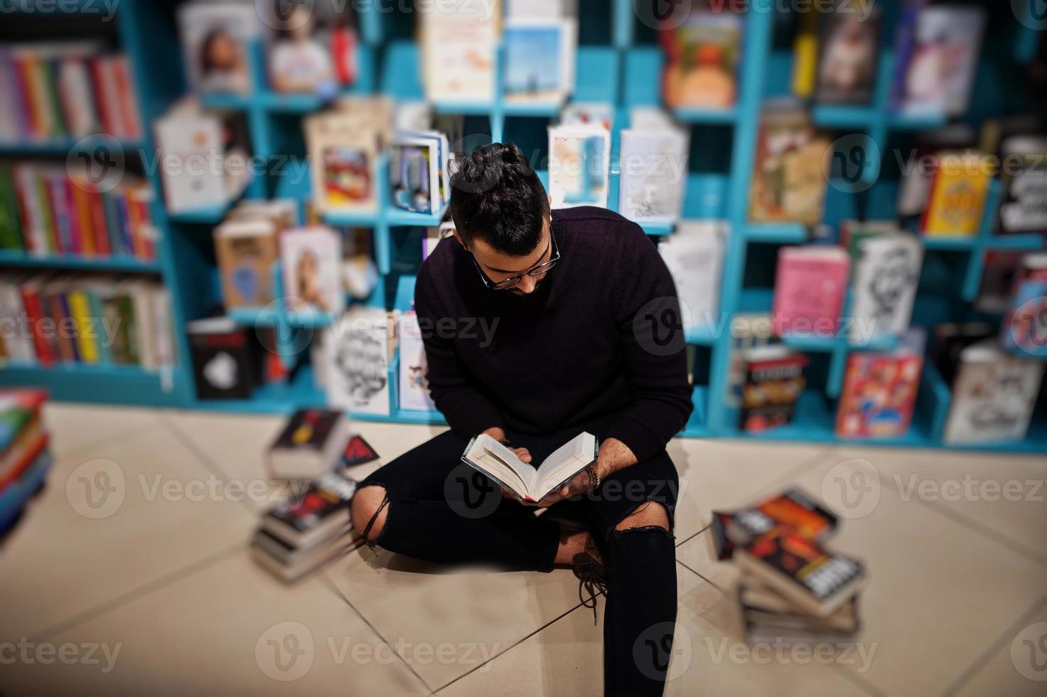 homem alto e inteligente estudante árabe, use gola rulê violeta e óculos, na biblioteca, sentado contra as prateleiras de livros. foto