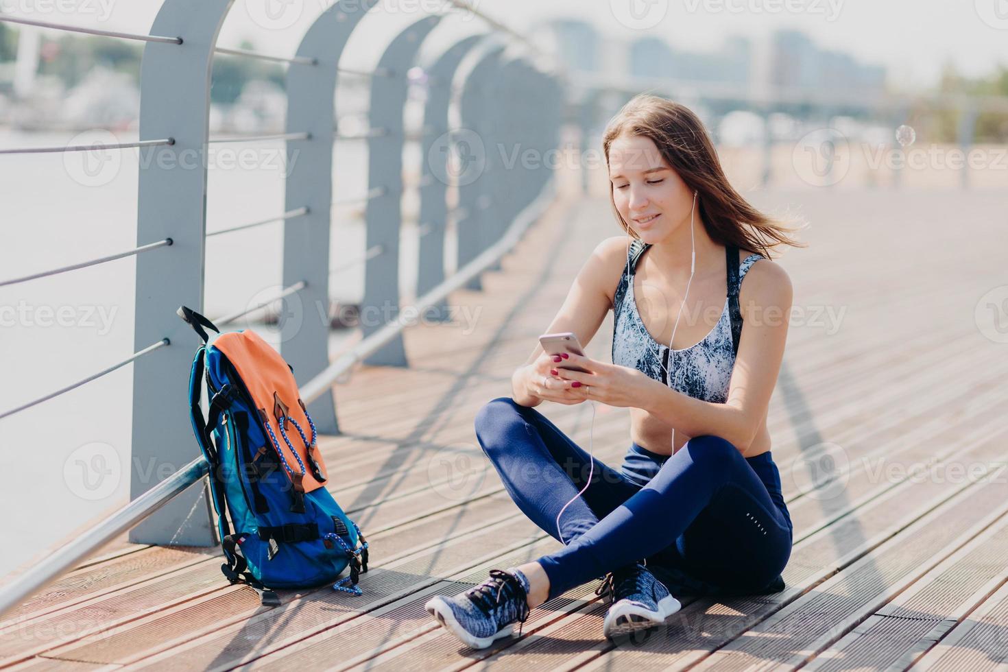 foto ao ar livre de mulher bonita encantada senta as pernas cruzadas, usa telefone celular moderno para ouvir música e mensagens com amigos, faz uma pausa após o exercício de corrida, usa conexão gratuita à Internet