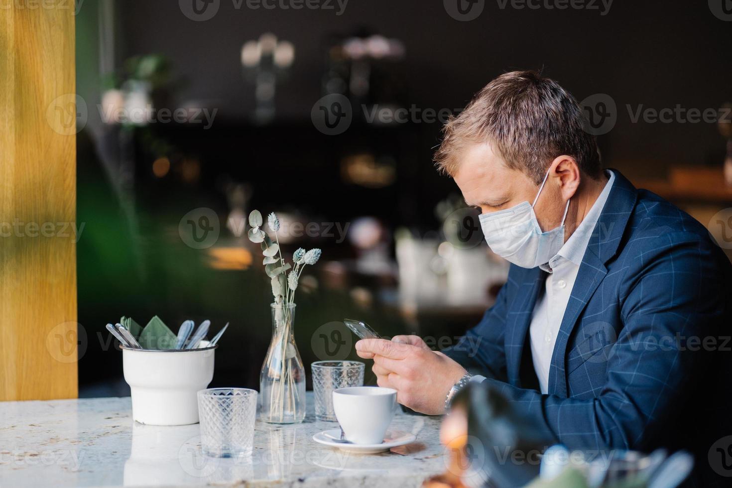 empresário masculino usa máscara médica durante surto de cornovírus, senta-se no café, espera pelo parceiro de negócios, bebe café, verifica o feed de notícias, conectado à internet sem fio. conceito de saúde foto