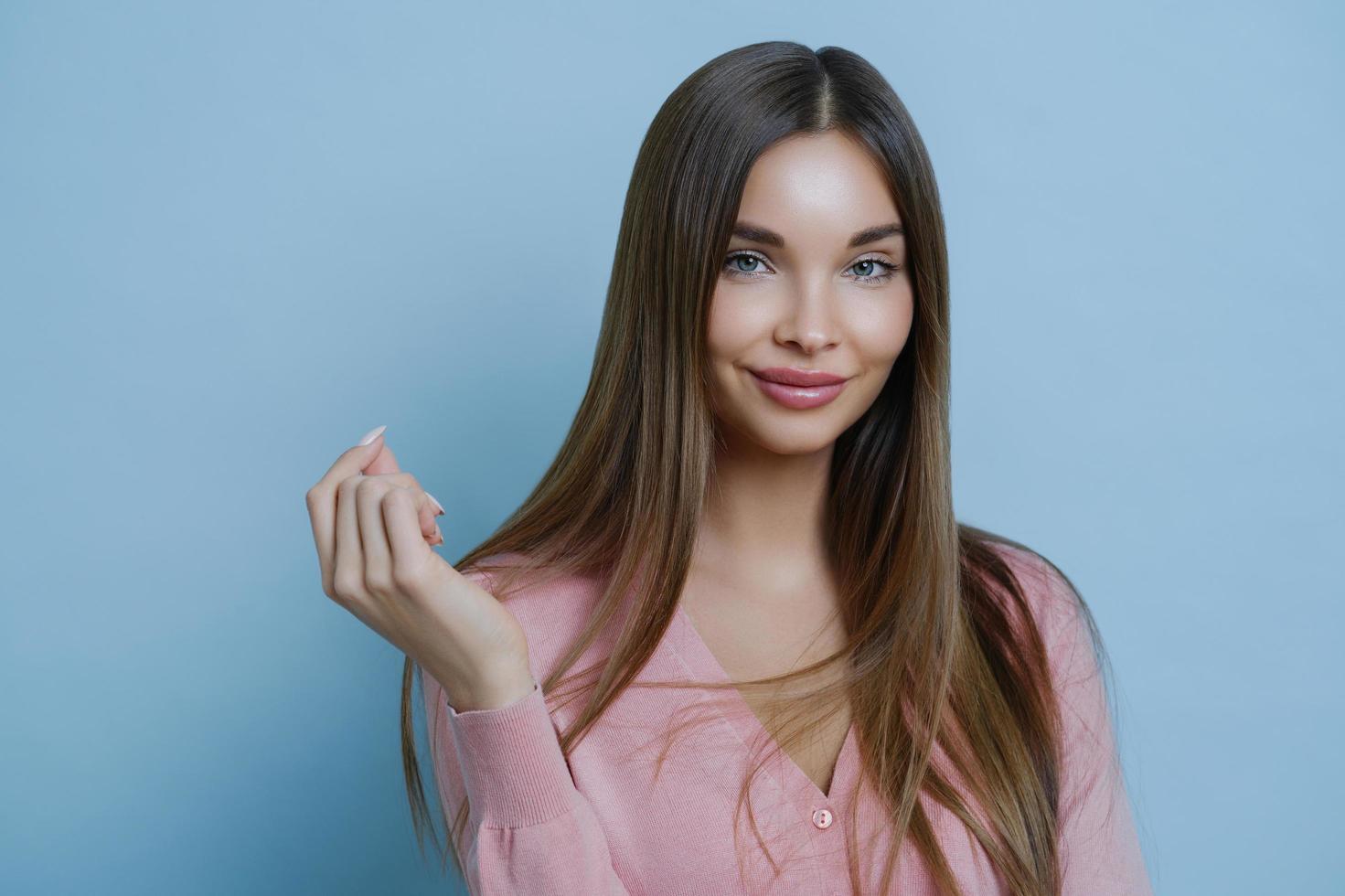 bela jovem europeia sem manchas e maquiagem, tem um visual fofo, mantém a mão levantada, sente-se encantada, posa contra um fundo azul, usa jumper rosado, vai participar de desfile de moda foto