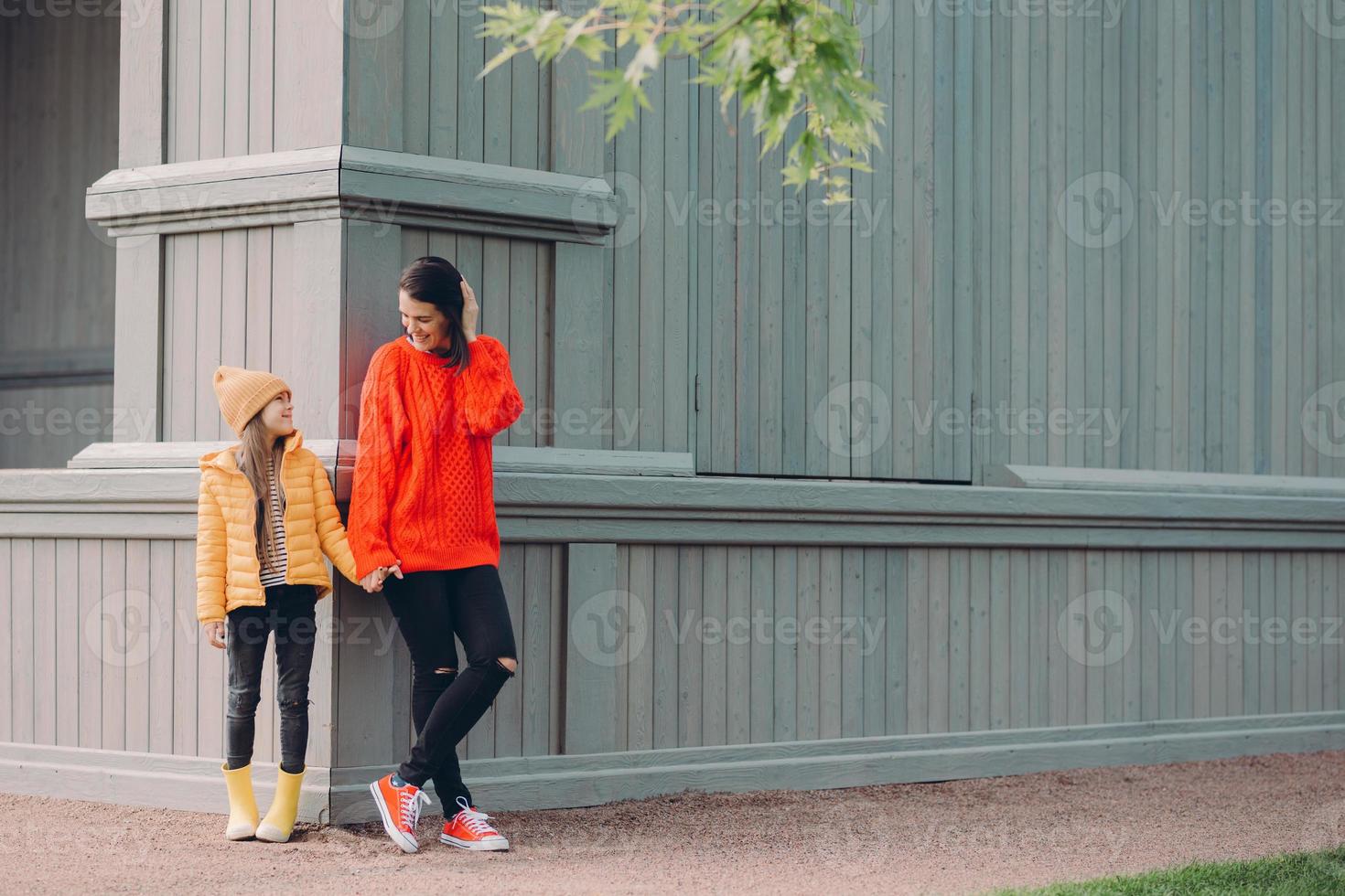 visão horizontal da mulher europeia elegante em suéter solto vermelho, jeans rasgado, de mãos dadas com sua irmã mais nova, posar fora juntos, caminhar ao ar livre, ter relações amigáveis foto