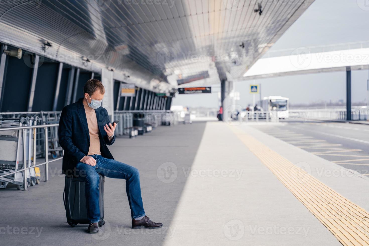 homem europeu senta-se na mala na estação da plataforma de ônibus, tenta encontrar o horário de transporte on-line no smartphone, aguarda o transporte, viaja durante uma situação de pandemia perigosa, usa máscara médica foto