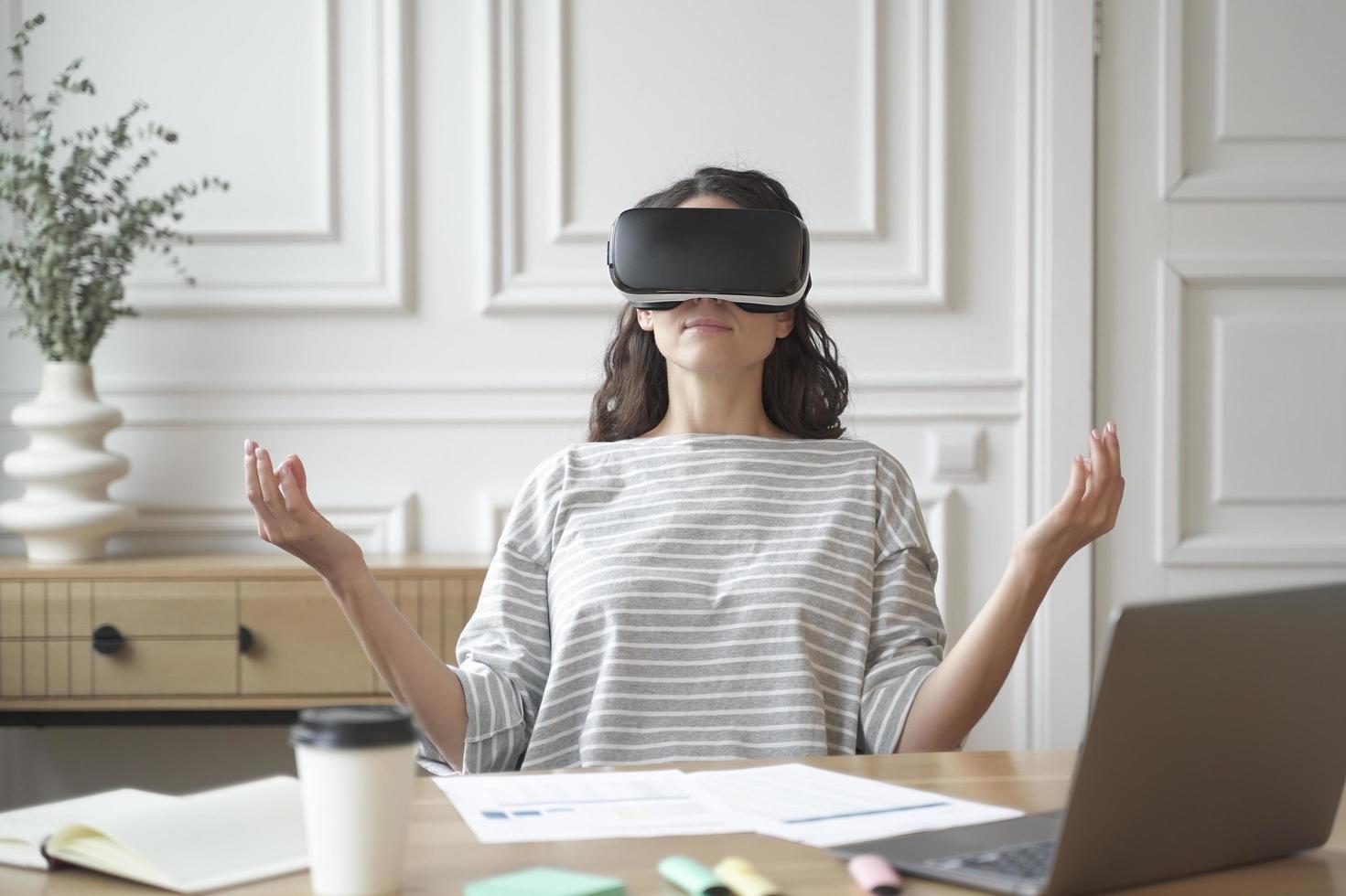 empregado de mulher pacífica meditando em óculos de realidade virtual, sentado em pose zen no local de trabalho foto