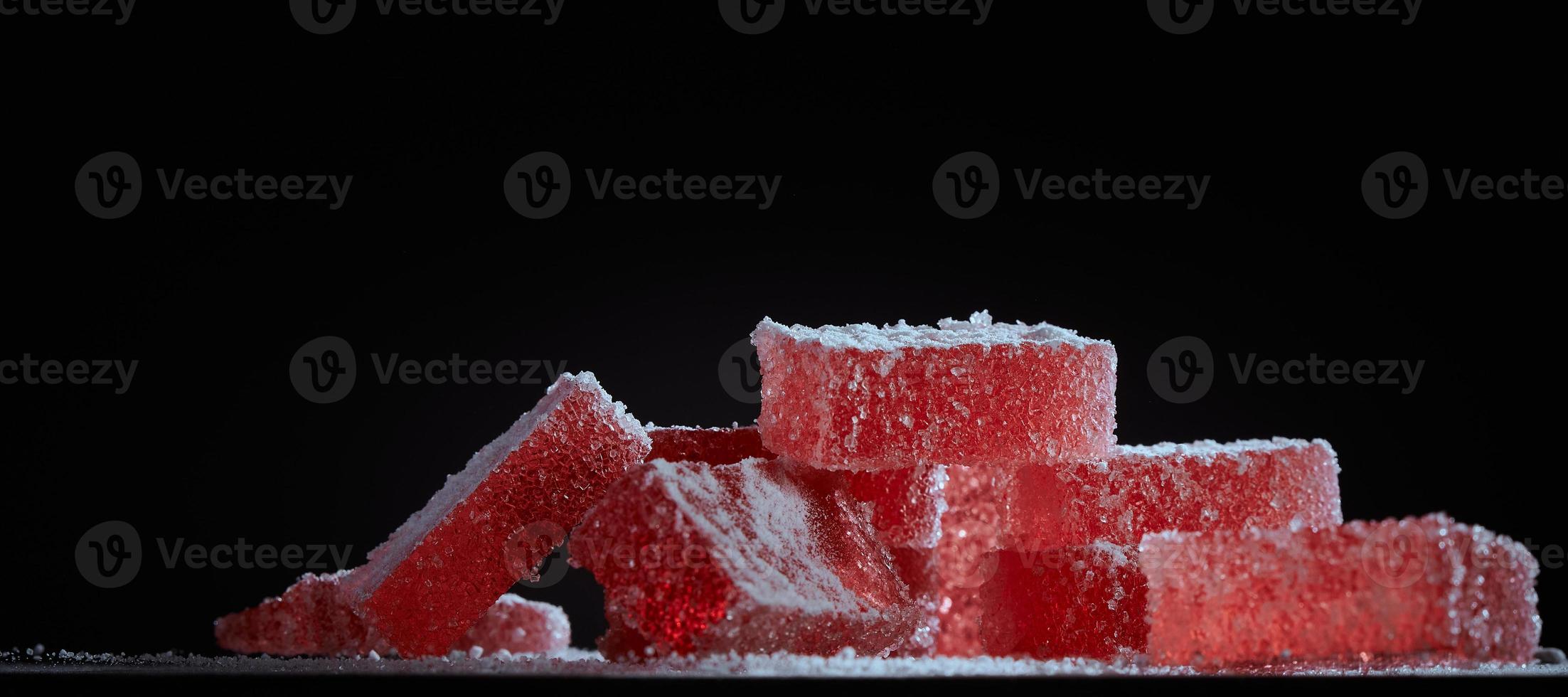 doces de geléia de marmelada vermelha brilhante em fundo preto. marmelada de sobremesa em forma de retângulos polvilhados com açúcar. a doçura do doce de geléia de baga. foto com espaço de cópia.