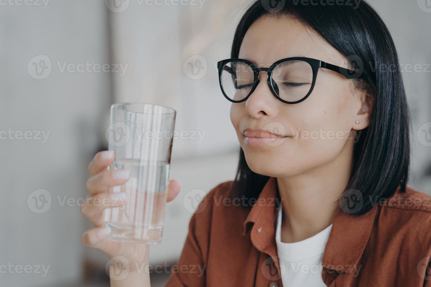 mulher feliz de óculos detém copo de água pura, olhos fechados. estilo de vida saudável, bem-estar foto