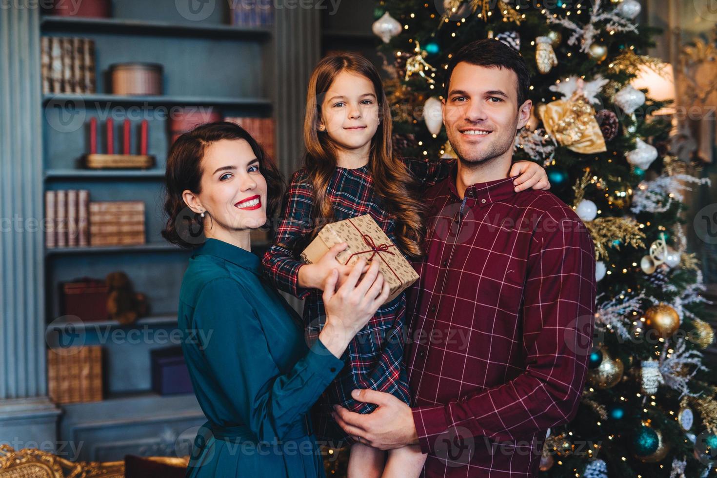 família feliz fica junto contra abeto decorado, olha alegremente para a câmera, segura o presente, comemora o ano novo no círculo familiar. menina feliz em passar férias com a mãe e o pai foto