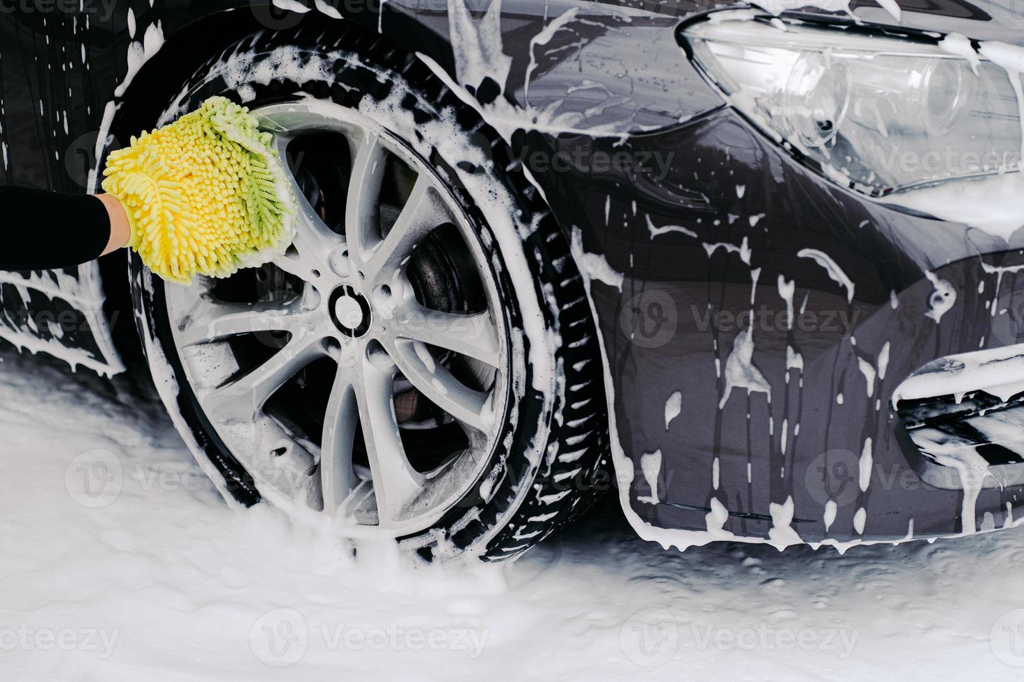 foto recortada de automóvel preto lavado pelo trabalhador de serviço no lava-jato. veículo coberto com bolhas de espuma. foco na roda.