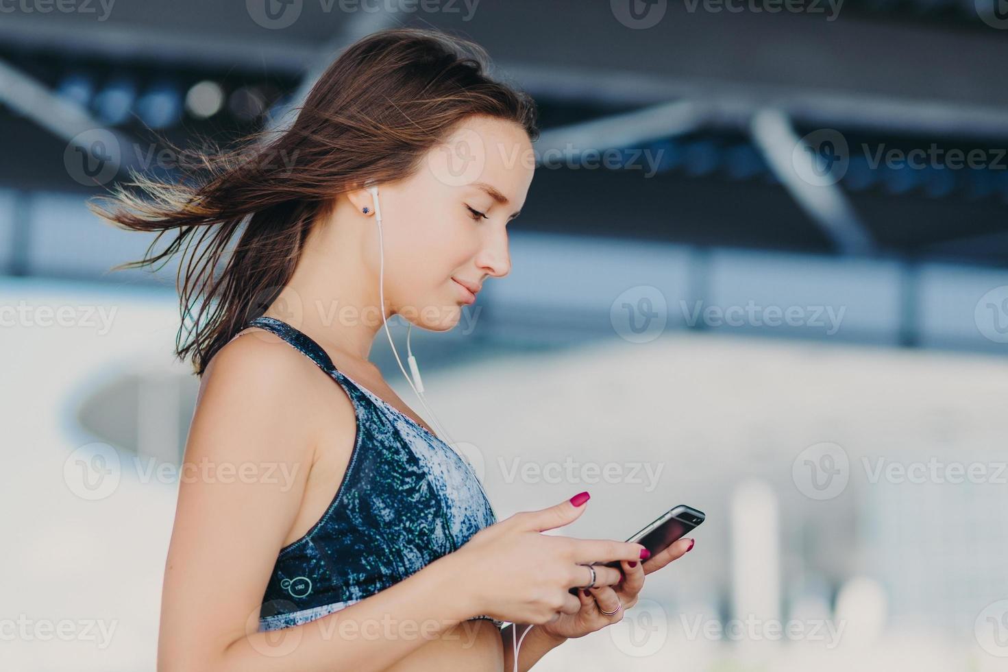 foto lateral de mulher adorável desportiva com cabelo escuro, vestida com roupa casual, segura telefone inteligente moderno, verifica a notificação, ouve música com fones de ouvido, gosta de conexão de internet de alta velocidade