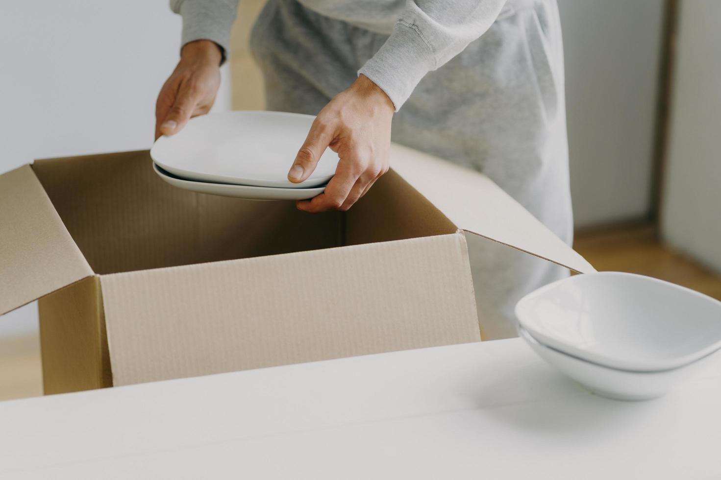 imagem recortada de homem ocupado sem rosto desconhecido desempacota caixas com pratos, segura pratos brancos, muda-se para um novo apartamento, arruma a cozinha, tem o dia da mudança. trazer novos pratos. conceito de realocação foto