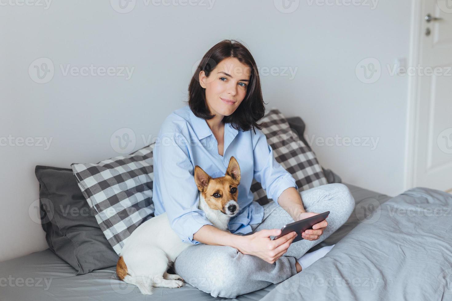 modelo feminino morena sonhadora de pijama senta as pernas cruzadas na cama confortável no quarto, segura o tablet moderno, abraça seu animal de estimação, sonha em caminhar ao ar livre. pessoas, descanso, tecnologia foto