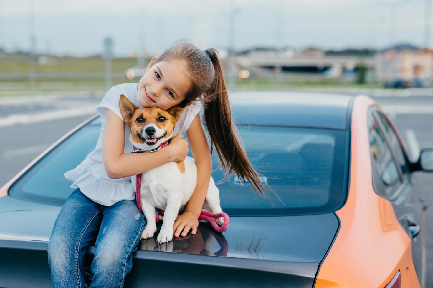 foto de criança feliz do sexo feminino tem rabo de cavalo abraça com amor seu animal de estimação favorito, posar no porta-malas do automóvel, brincar juntos, caminhar ao ar livre, desfrutar da união. conceito de crianças e animais