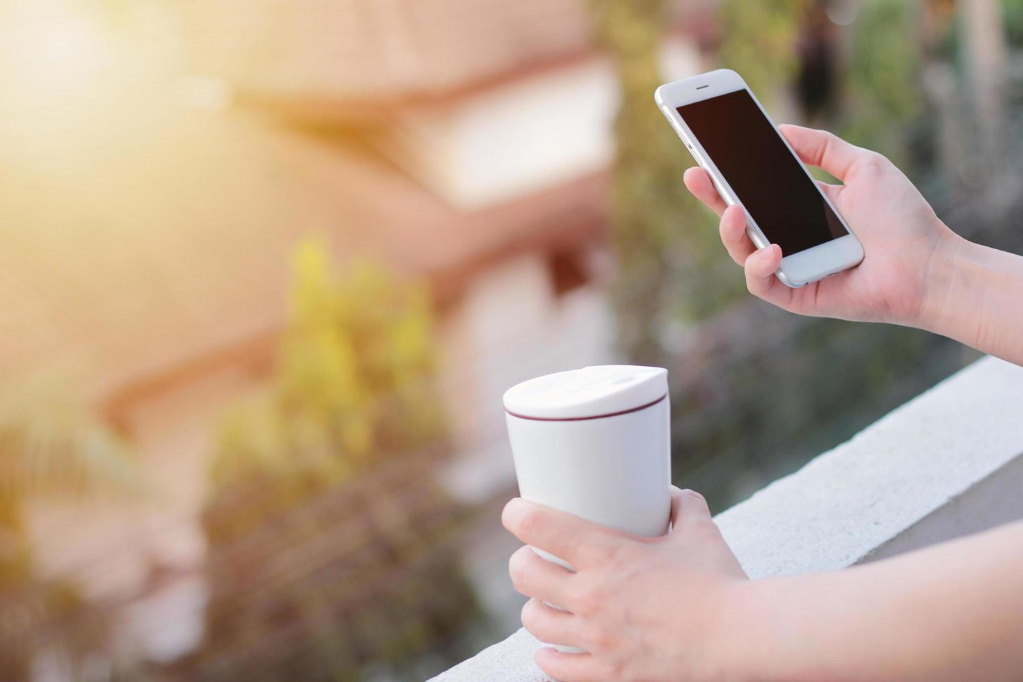 close-up de mãos de mulher usando smartphone e segurando a caneca térmica, copie o espaço com luz solar foto