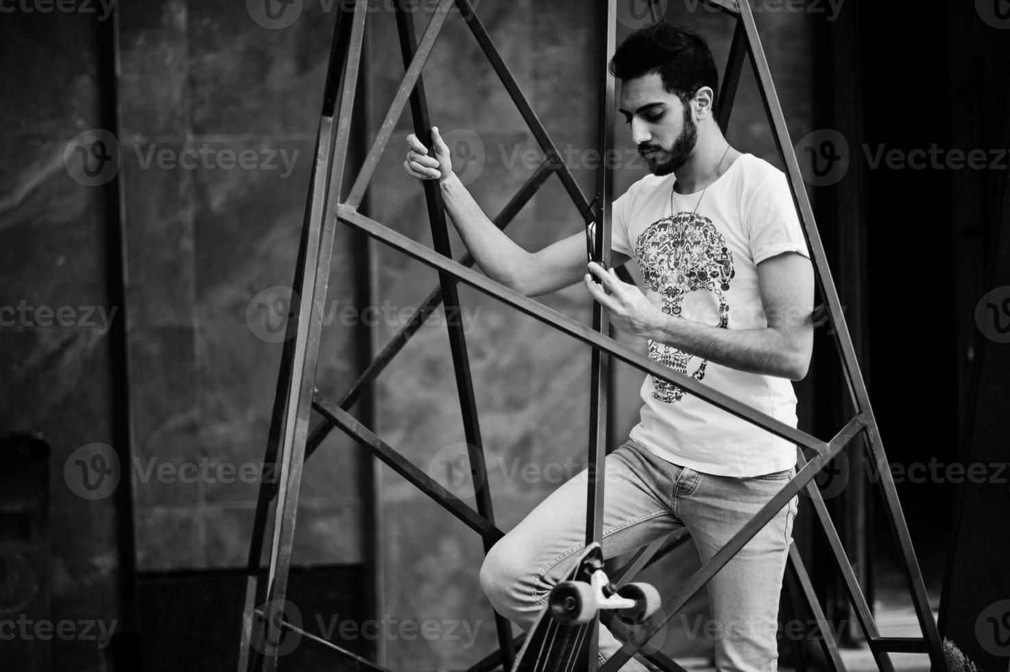 homem árabe de estilo de rua em óculos com longboard posou dentro da construção da pirâmide de metal. foto