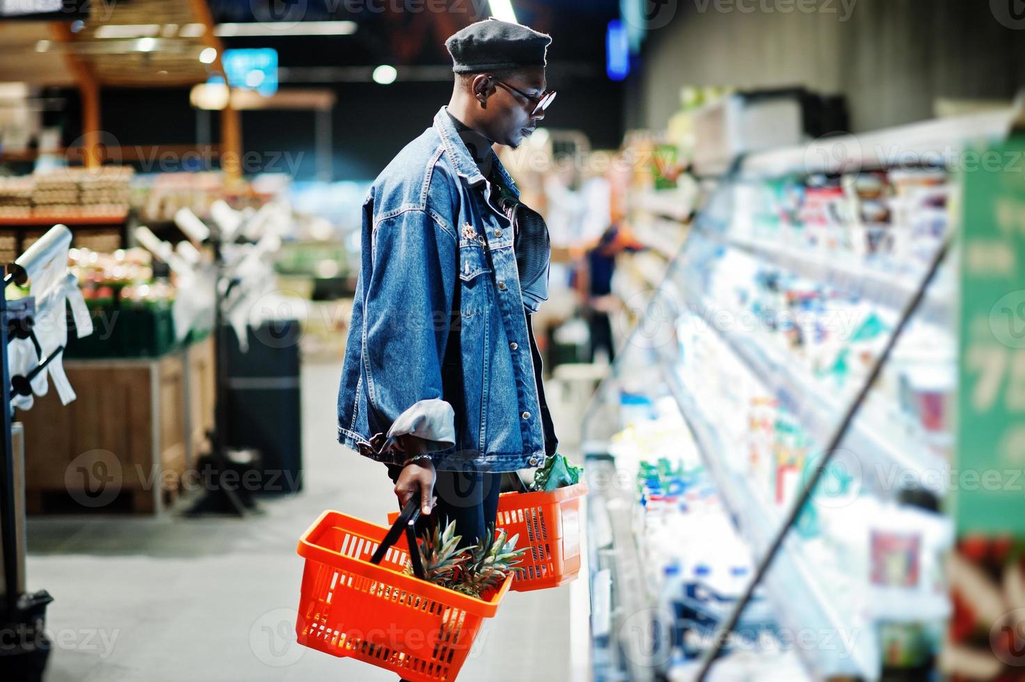 homem afro-americano casual elegante na jaqueta jeans e boina preta segurando duas cestas, perto da geladeira e compras no supermercado. foto