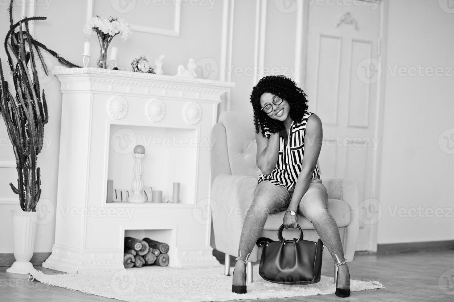 linda mulher afro-americana com cabelo afro encaracolado e óculos, bolsa posada no quarto, sentado na cadeira amarela. foto