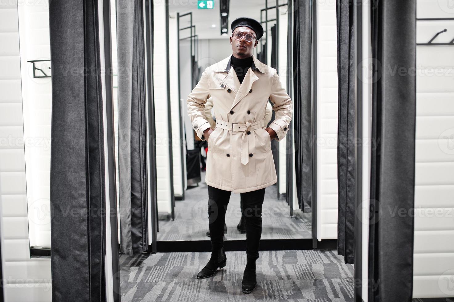 homem afro-americano casual elegante na boina preta e capa de chuva branca na loja de roupas de provador. foto