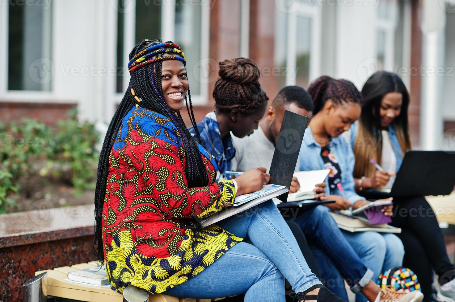 grupo de cinco estudantes universitários africanos passando tempo juntos no campus no pátio da universidade. amigos afro negros estudando no banco com itens escolares, notebooks laptops. foto