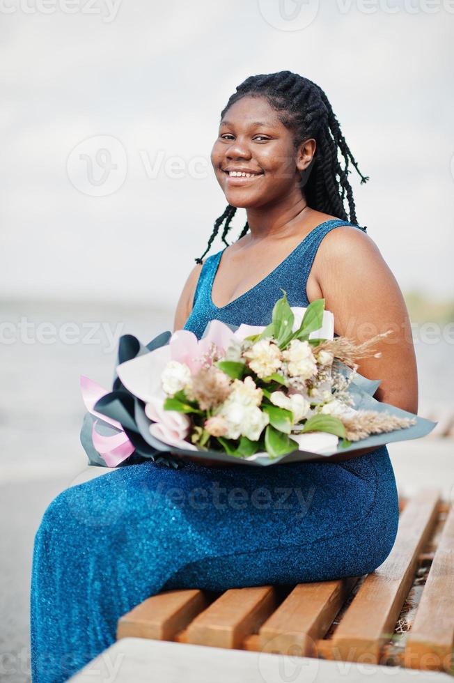 modelo afro-americano de pele escura plus size posou em um vestido azul brilhante com buquê de flores sentado no banco contra o lado do mar. foto