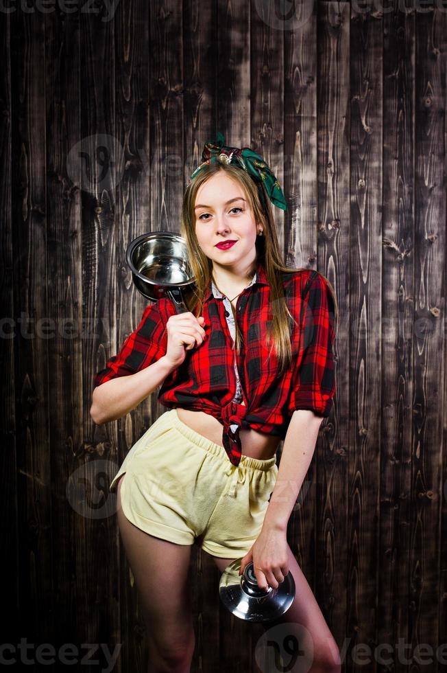 jovem dona de casa engraçada na camisa quadriculada e short amarelo pin up estilo com panela em fundo de madeira. foto