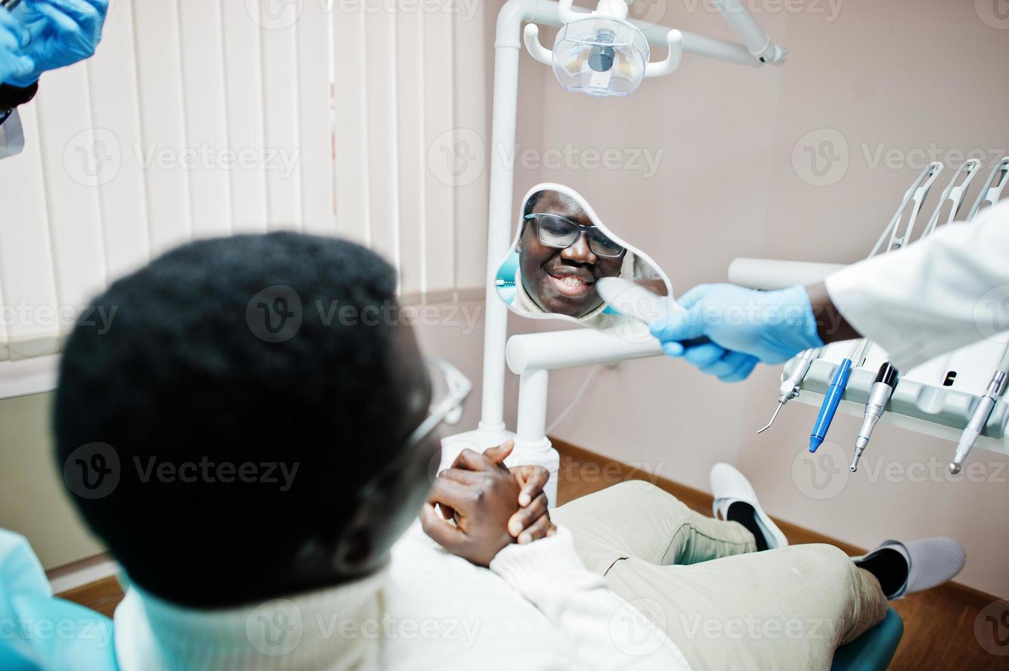 paciente homem afro-americano na cadeira odontológica. consultório de dentista e conceito de prática médica. dentista profissional ajudando seu paciente na odontologia médica e mostrando a ele um espelho. foto