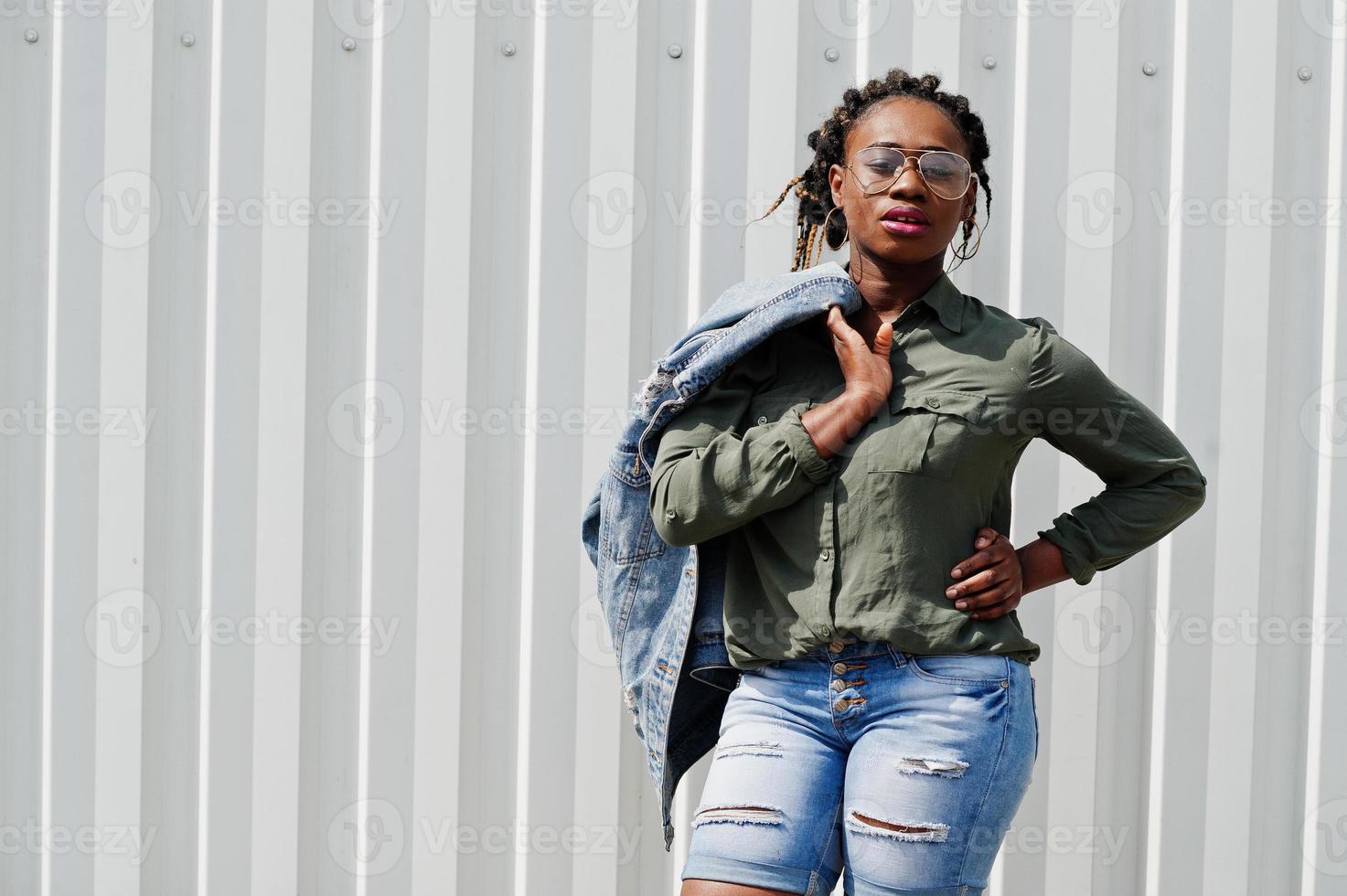 mulher africana com cabelo de dreads, jaqueta jeans e shorts, usa óculos posados contra a parede de aço branca. foto