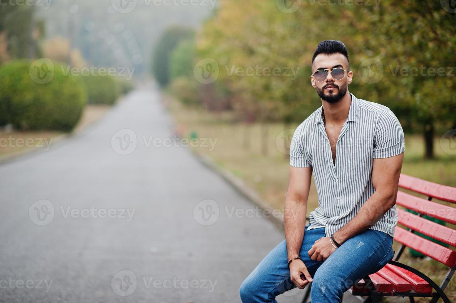 homem de barba árabe alto na moda usar camisa, jeans e óculos de sol posou no parque no banco. foto