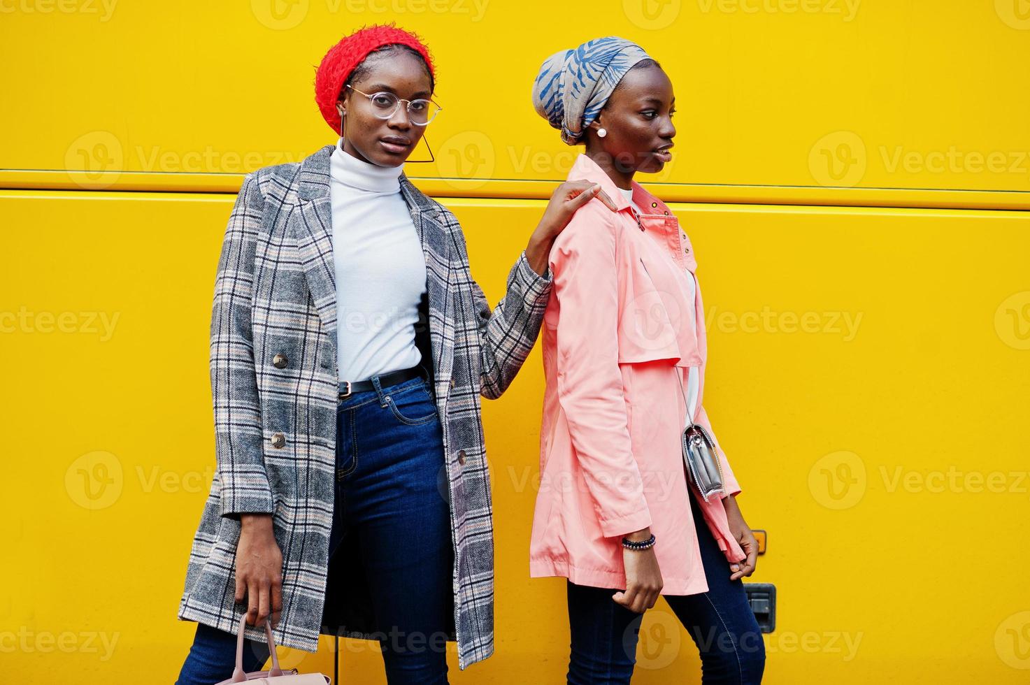 duas jovens mulheres muçulmanas africanas elegantes, atraentes, altas e magras em hijab ou lenço de cabeça de turbante e casaco posaram contra ônibus amarelo. foto