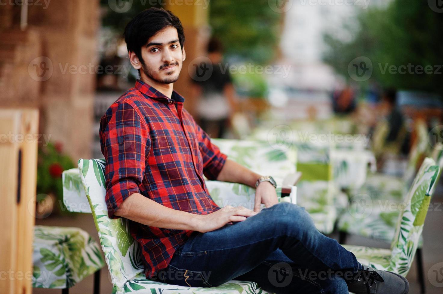 homem jovem estudante indiano na camisa xadrez vermelha e jeans sentado no café à noite. foto