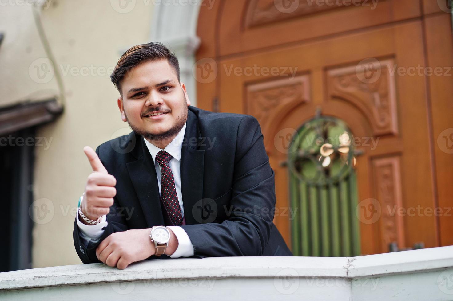 empresário indiano elegante com roupa formal, apoiando-se em uma grade contra a porta no centro de negócios. ele homem feliz e mostra o polegar para cima. foto