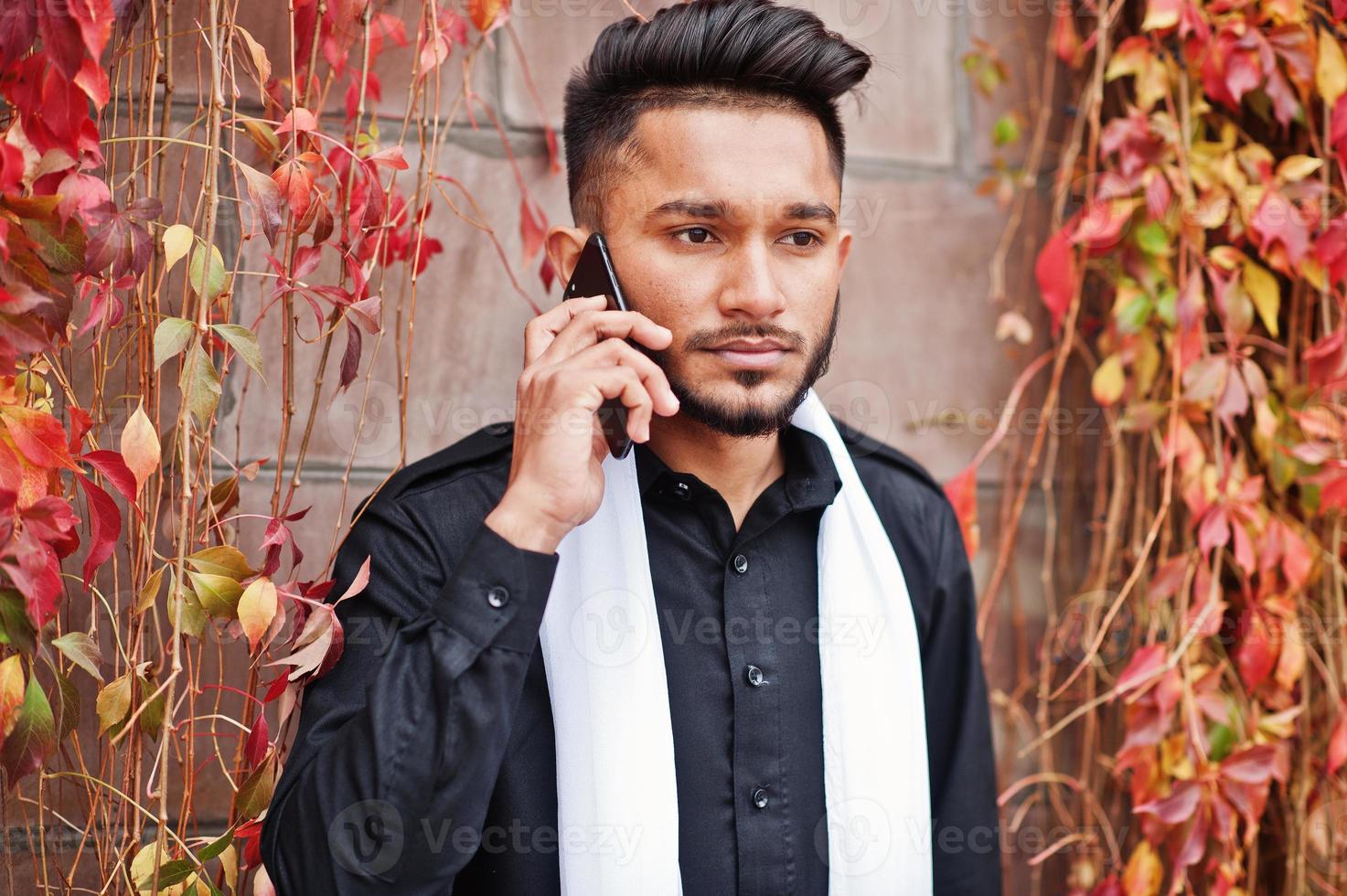 homem elegante indiano em roupas tradicionais pretas com lenço branco posou ao ar livre contra a parede de folhas vermelhas falando no celular. foto
