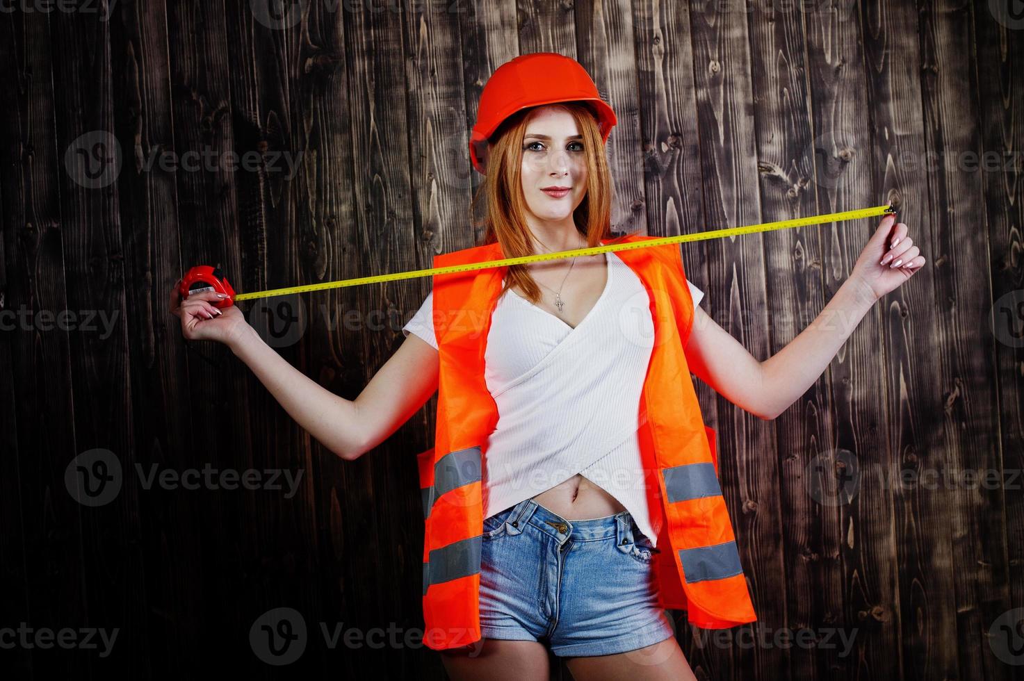 mulher engenheira em laranja proteger o capacete e a jaqueta de construção contra o fundo de madeira, segurando a fita métrica. foto