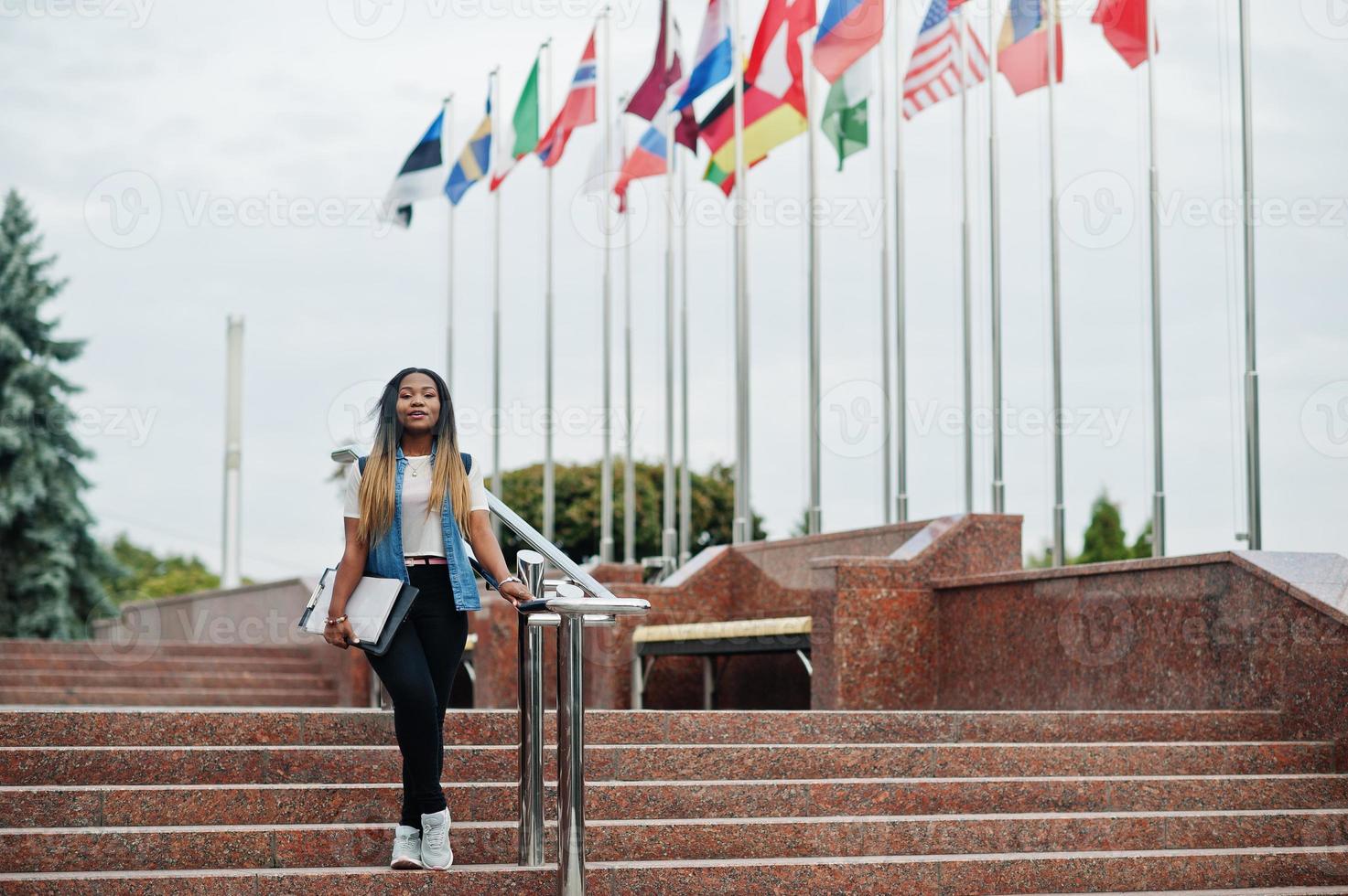 mulher estudante africana posou com mochila e itens escolares no pátio da universidade, contra bandeiras de diferentes países. foto