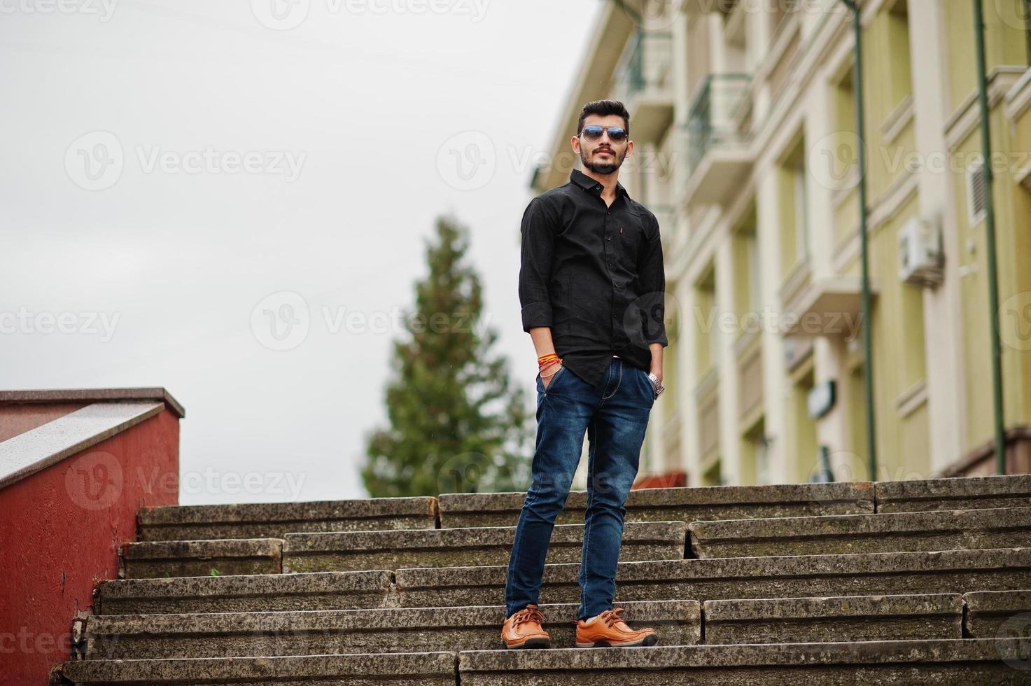 homem elegante indiano na camisa preta e óculos de sol posou ao ar livre. foto