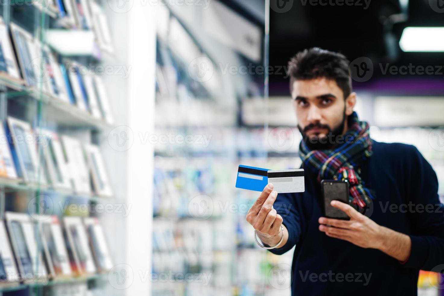 comprador de cliente de homem de barba indiana na loja de celular escolhe seu novo smartphone e segurando cartões de crédito. conceito de povos e tecnologias do sul da Ásia. loja de celulares. foto