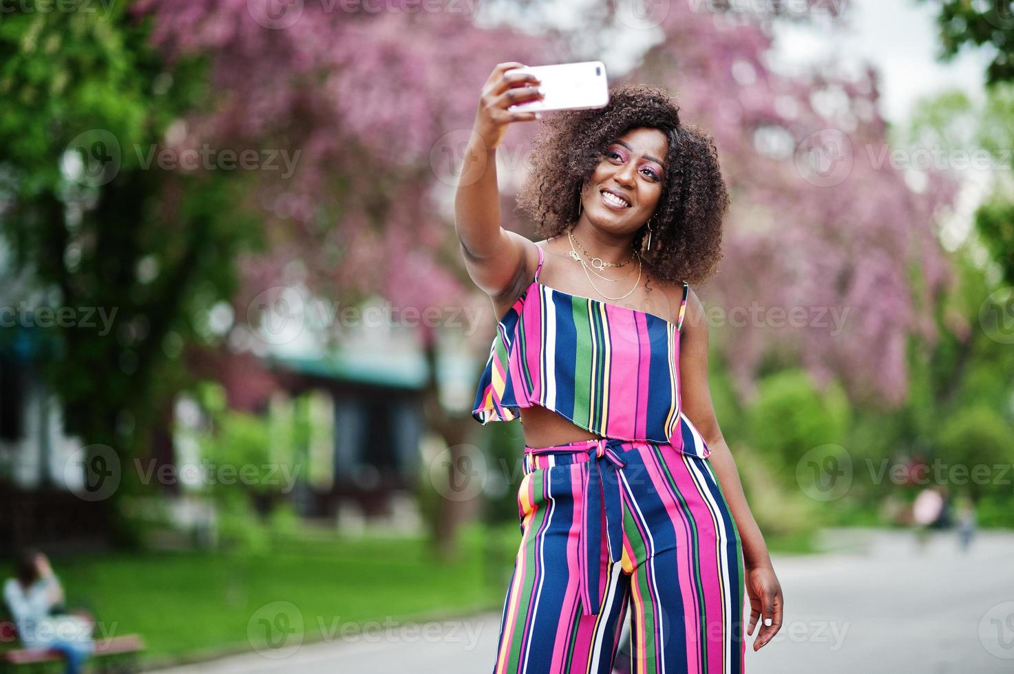 mulher afro-americana elegante em macacão listrado rosa posou na rua da flor da primavera e fazendo selfie pelo telefone celular. foto