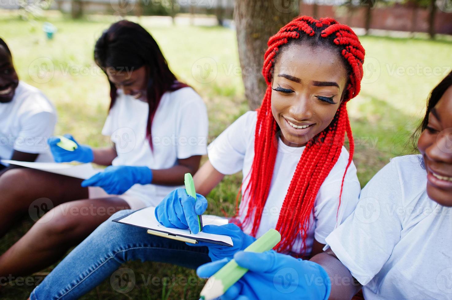 grupo de voluntários africanos felizes sentados debaixo da árvore no parque e escrever algo nas pranchetas. áfrica voluntariado, caridade, pessoas e conceito de ecologia. foto