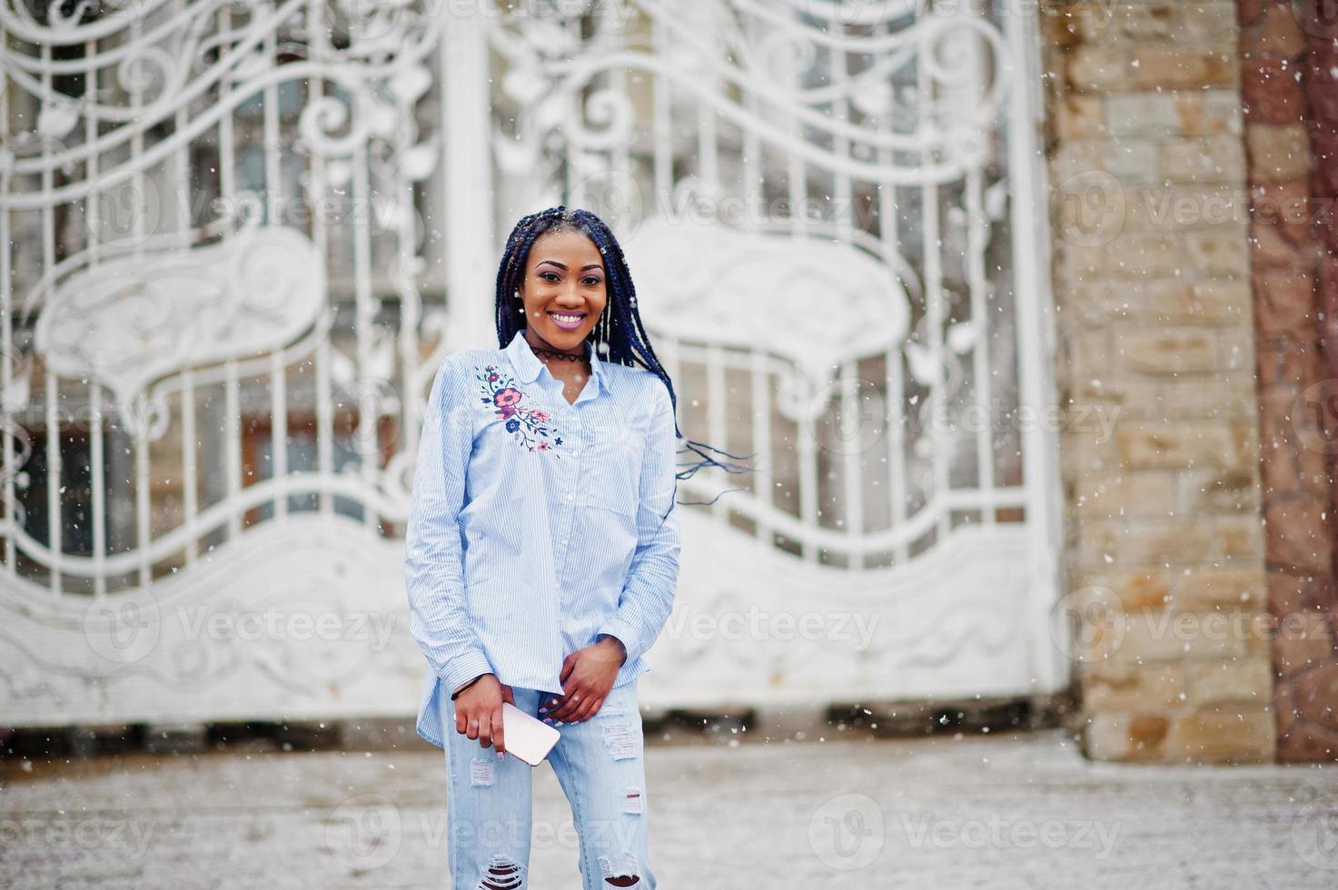 menina afro-americana elegante com dreads segurando o celular na mão, ao ar livre contra portões reais brancos em tempo de neve. foto