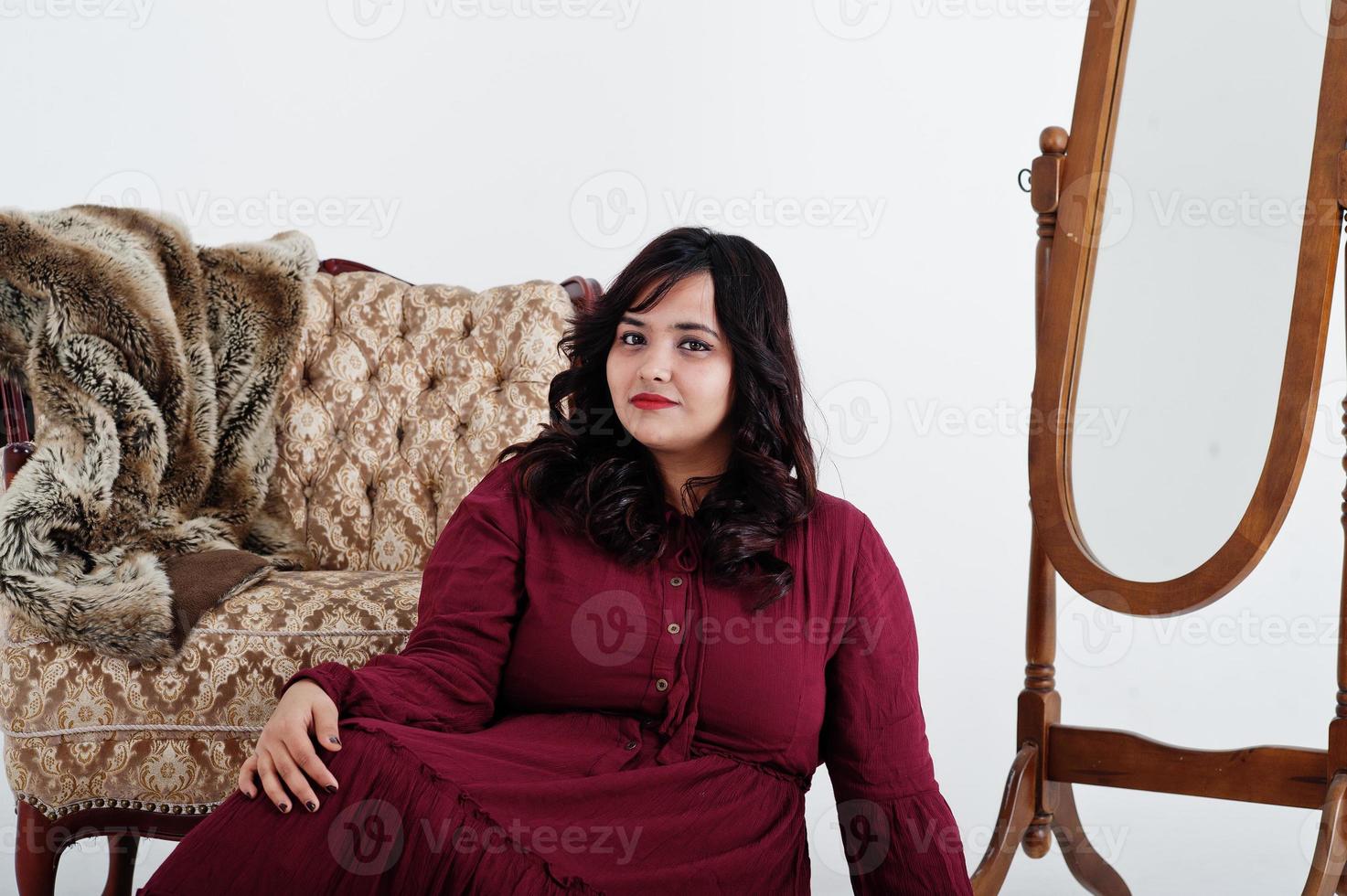 mulher atraente do sul da Ásia em vestido vermelho escuro posou no estúdio em fundo branco contra espelho e cadeira. foto