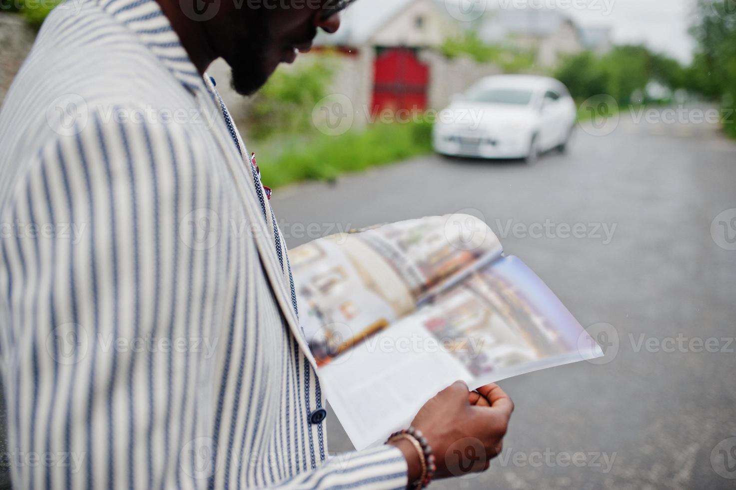 homem afro-americano rico e elegante de blazer e óculos lê revista contra seu carro. foto