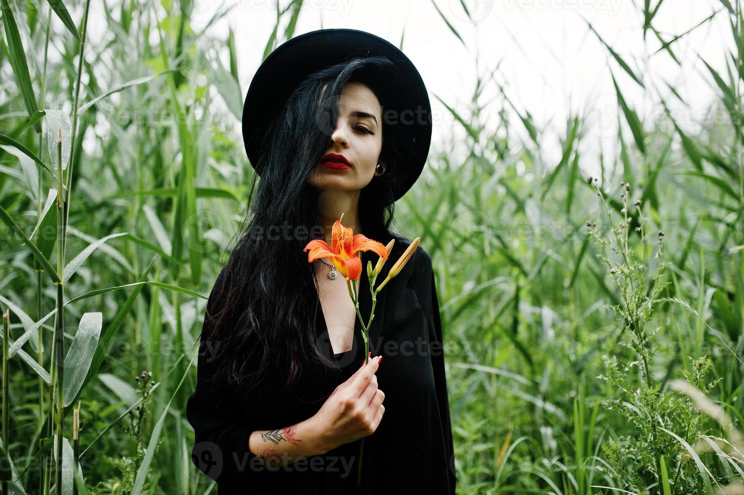 garota sensual toda em preto, lábios vermelhos e chapéu. mulher dramática gótica segura flor de lírio laranja na cana comum. foto