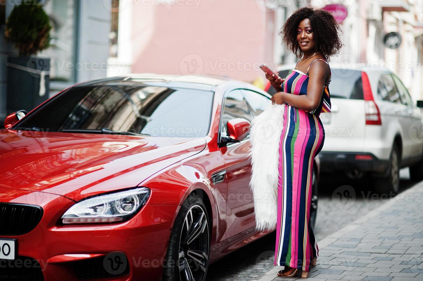 mulher afro-americana elegante em macacão listrado rosa com casaco de pele falso fofo posou contra carro de luxo vermelho rico com telefone celular na mão. foto