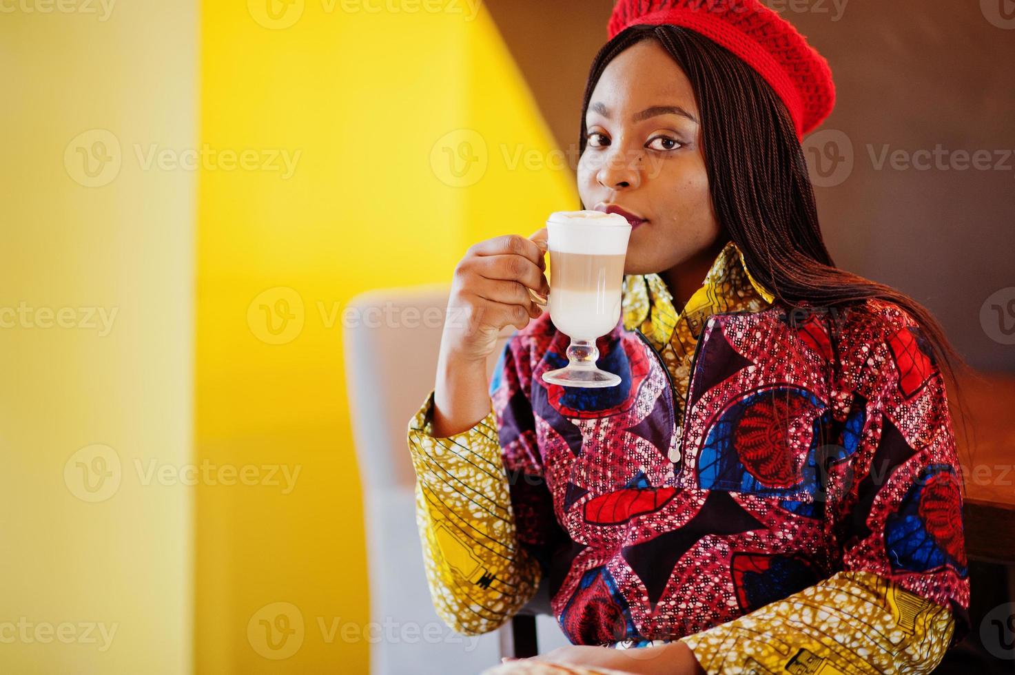mulher afro-americana entusiasmada com roupa colorida na moda com boina vermelha relaxando no café aconchegante com uma xícara de café com leite quente nas mãos. foto