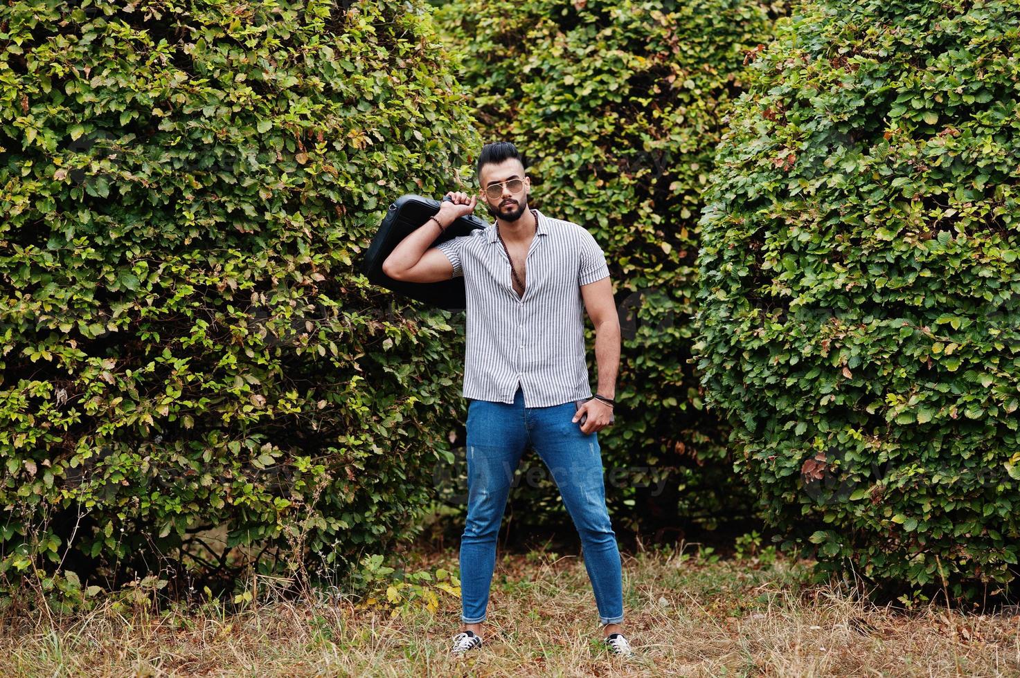 homem de barba árabe alto na moda usar camisa, jeans e óculos de sol posou no parque e segure a mala. foto