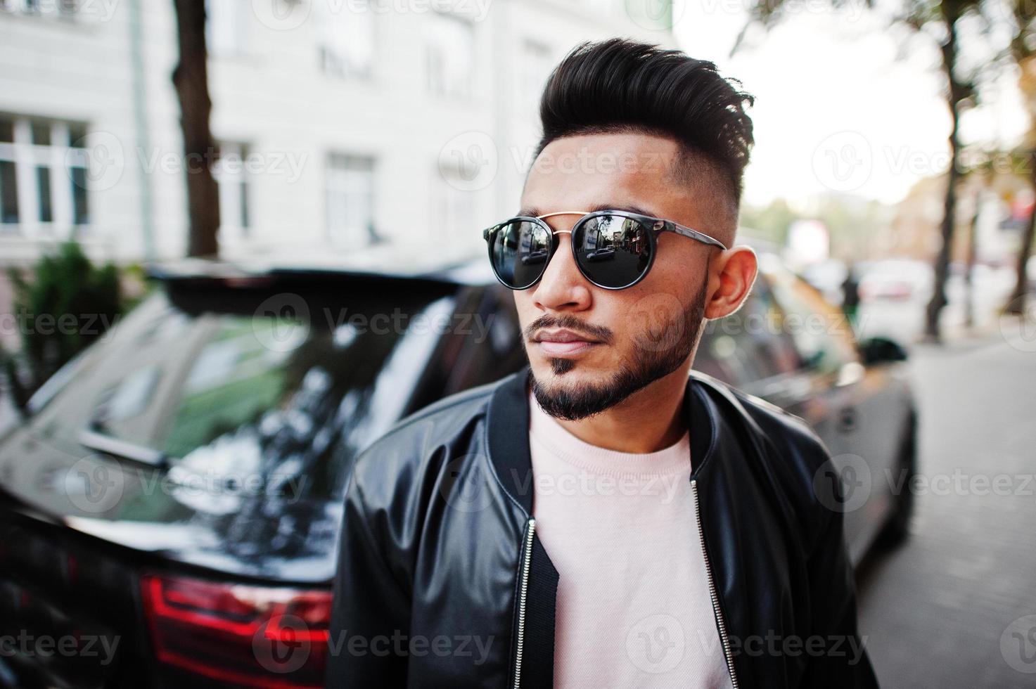 homem de barba indiana elegante na jaqueta de couro preta e óculos de sol contra carro suv de negócios. modelo da índia posou ao ar livre nas ruas da cidade. foto